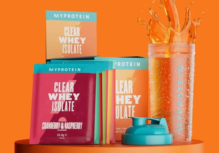 Opakowanie czystej serwatki na podium, prezentujące różne smaki, w tym żurawinę i maliny oraz pomarańczowe mango, obok shakera marki Myprotein.